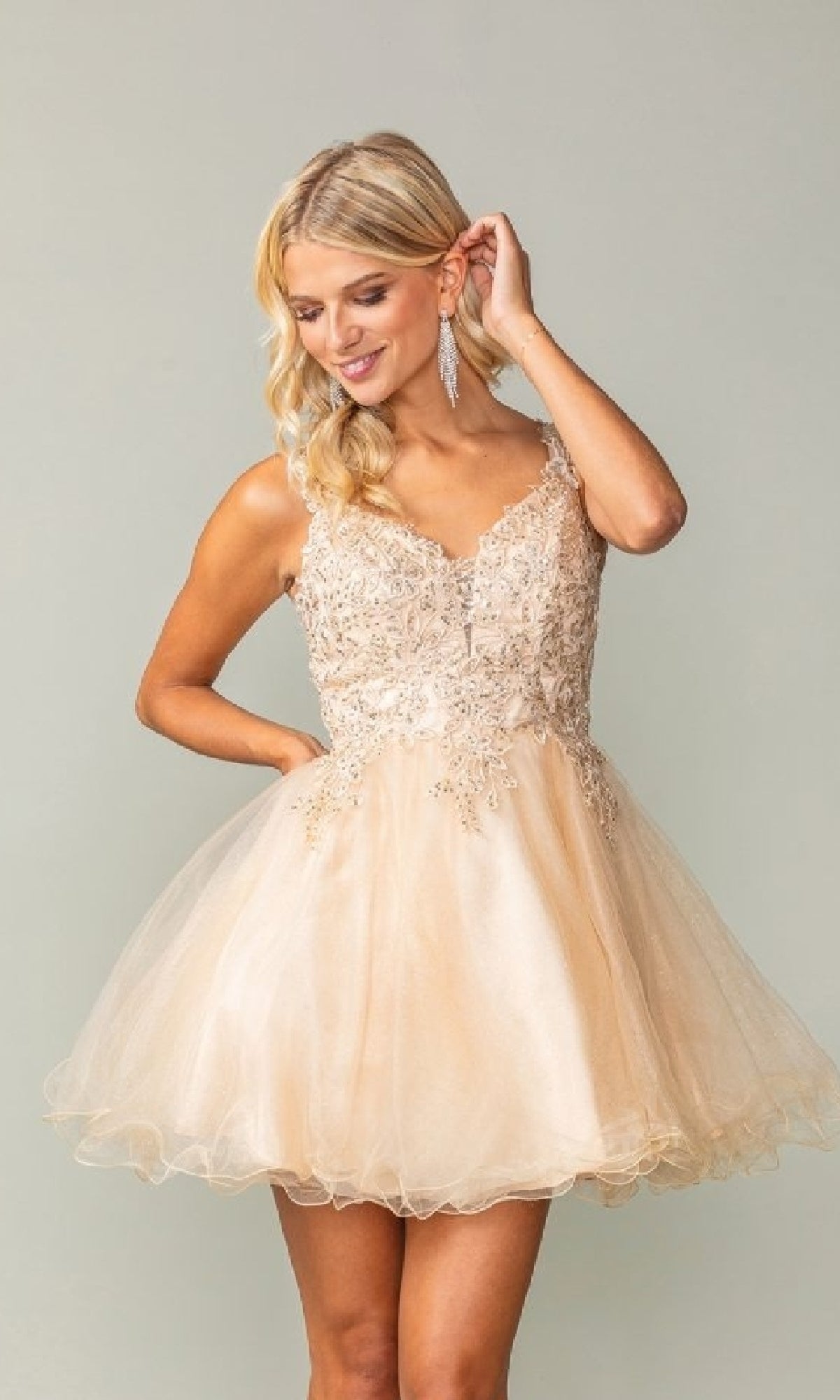 A woman poses in the Tulle Babydoll Homecoming Dress: Dancing Queen 3377, featuring a short, sleeveless champagne bodice with lace and sequin accents and a playful tulle skirt.